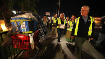 Photograph of several people wearing neon vests walking down a street at night; on the side there is a shopping cart filled with clothes and bags